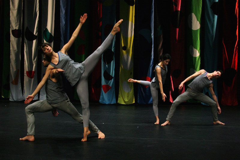 Compagnie CNDC-Angers dancers strike various angular poses. A female dancer in the foreground extends her left leg in the air as her upper body is tilted to the right